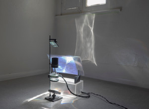 Apparatus on the ground in front of a window. Lenses are held by a vertical steel rod, and a rectangular lens sits on a semi-transparent box that is lit by sunlight. The various lenses cast patterns on the wall, some of which are white and others are more like rainbows.